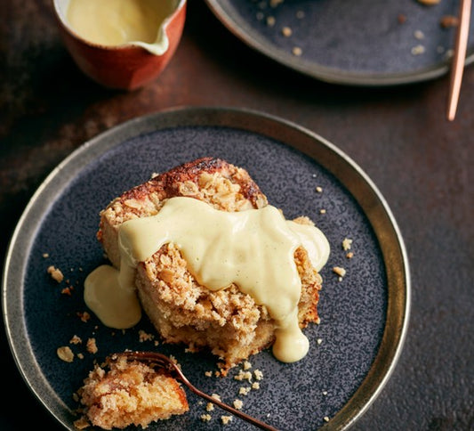 Close-up of a golden, crumbly apple crumble with cinnamon-spiced apples, topped with creamy custard being poured over it.